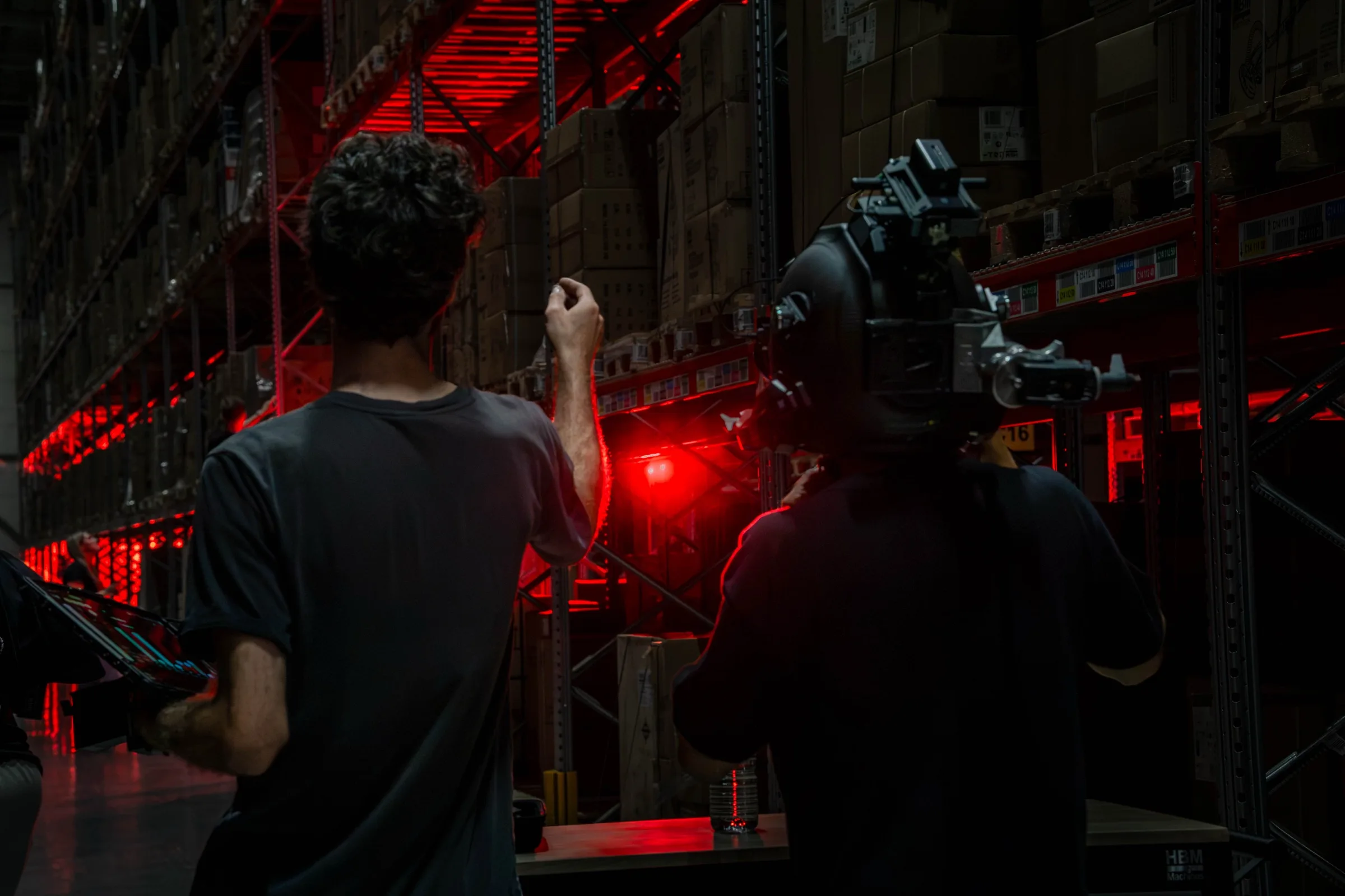 Een crew filmt tussen rood verlichte magazijnstellingen